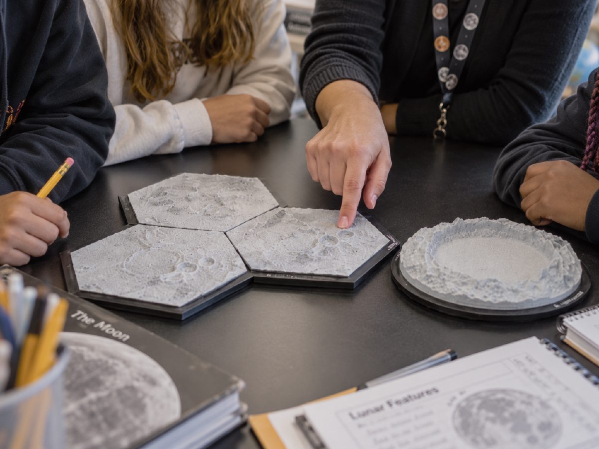 Teacher pointing at a crater on hexagonal lunar terrain tiles in a classroom